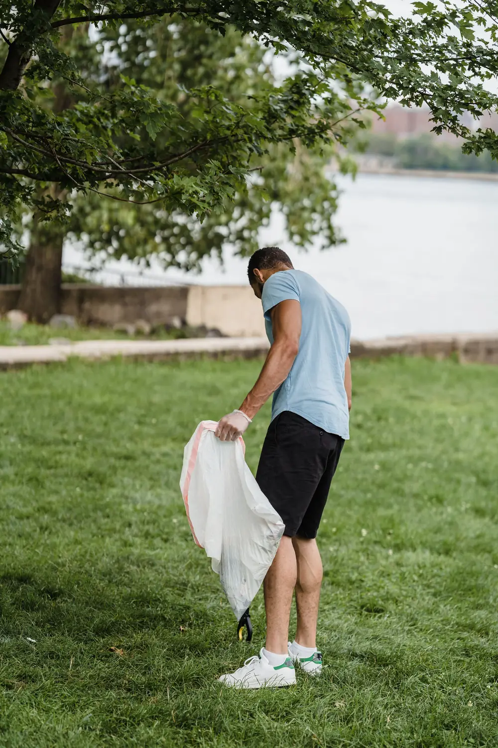 Ein junger Mann sammelt mit einem Müllsack Abfall auf einer Wiese am Fluss